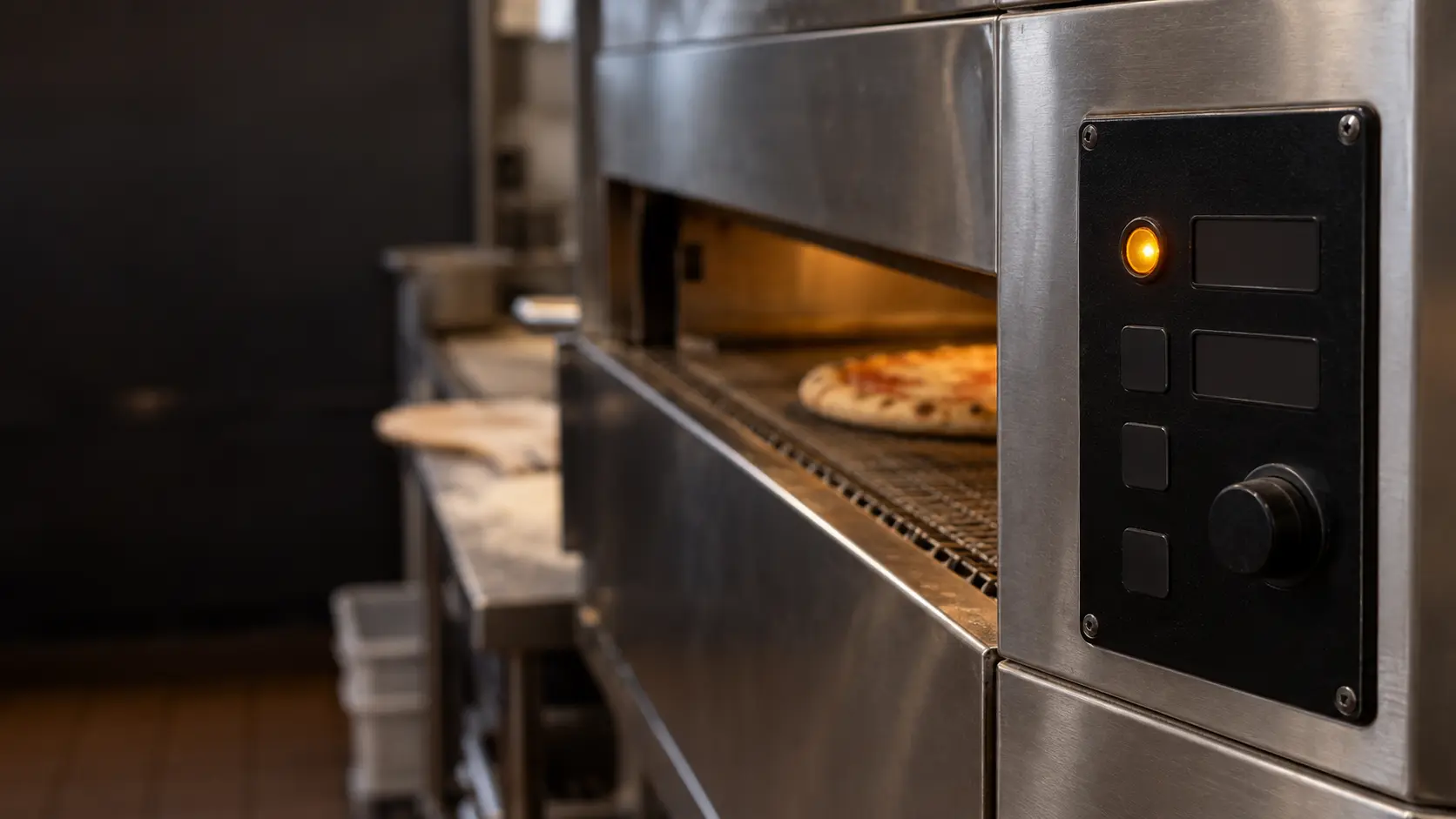 Close view of the dark digital control panel on the side of a stainless conveyor pizza oven, one abstract amber indicator glowing beside unlit segments, the edge of the pizza oven belt blurred behind it.