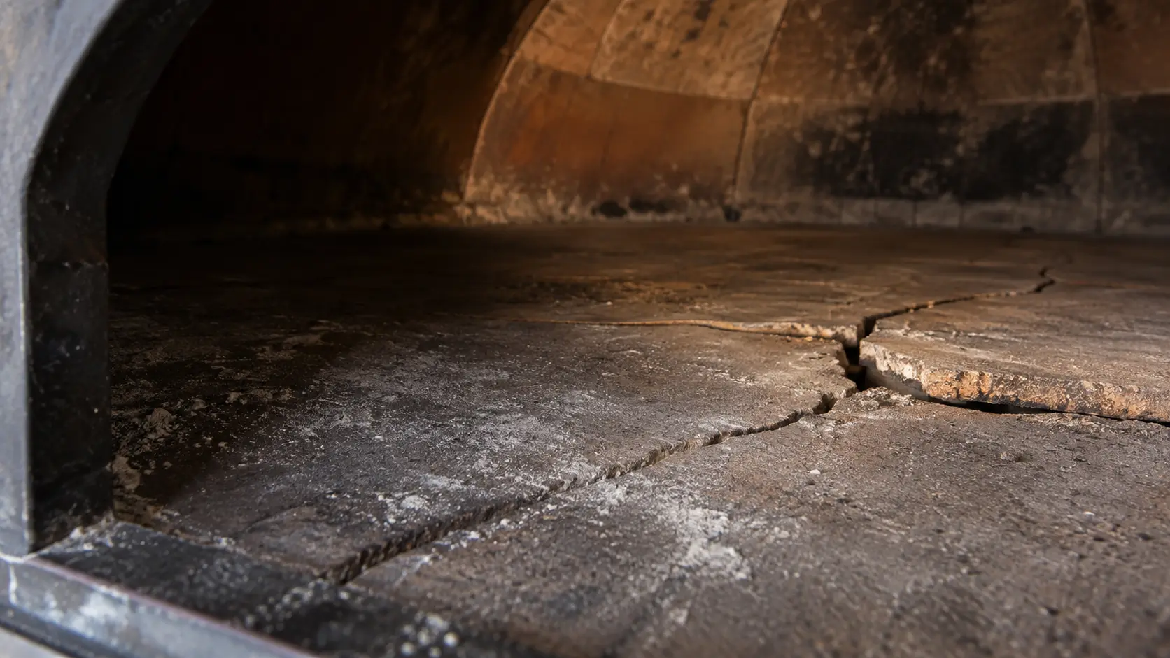 Close view of the cold stone floor inside a wood-fired pizza oven, a wide crack running across two cooking tiles with one edge lifted slightly above the other, a light dusting of flour and ash, the curve of the pizza oven dome at the top of the frame.