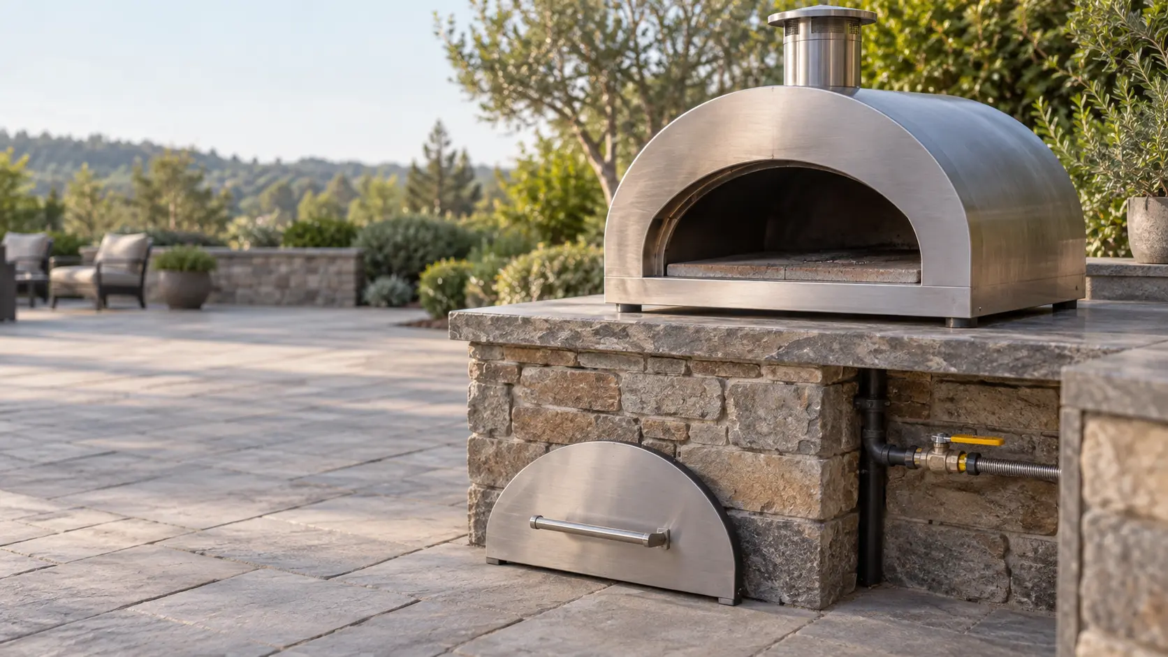 A cold stainless outdoor gas pizza oven standing alone on a stone patio counter in soft daylight, the pizza oven's door lifted off and set on the ground beside the counter, its dark empty stone floor visible through the open mouth, the gas shutoff valve on the line below turned closed, patio chairs pushed back.