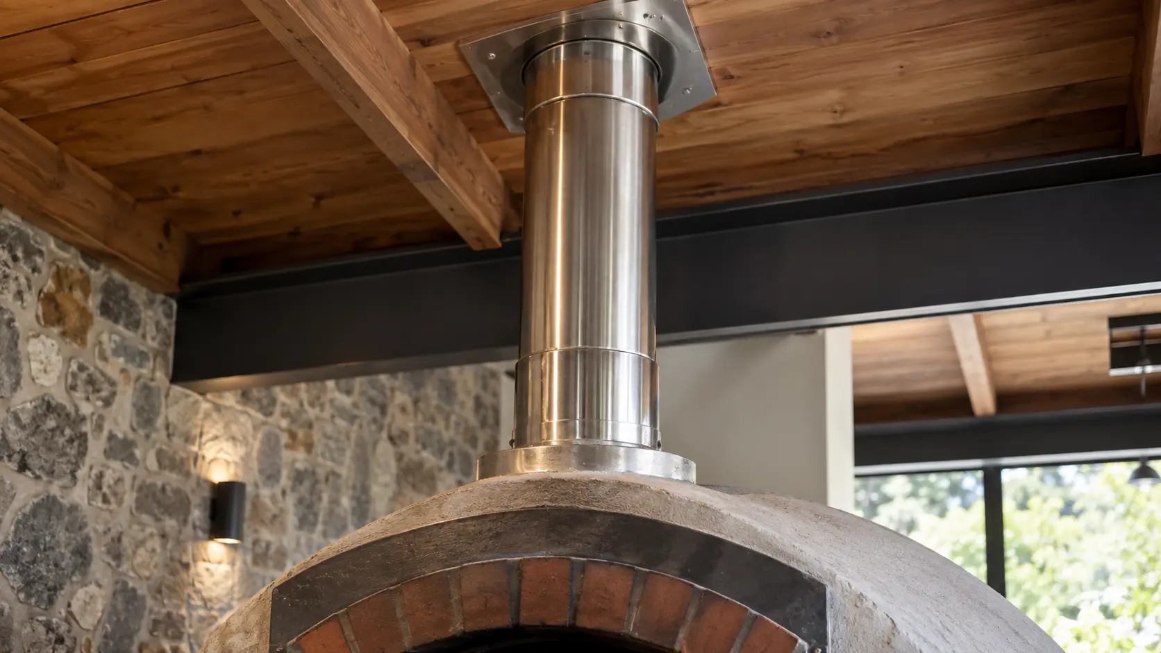 Close on a stainless flue pipe rising from a pizza oven toward a roof cap.